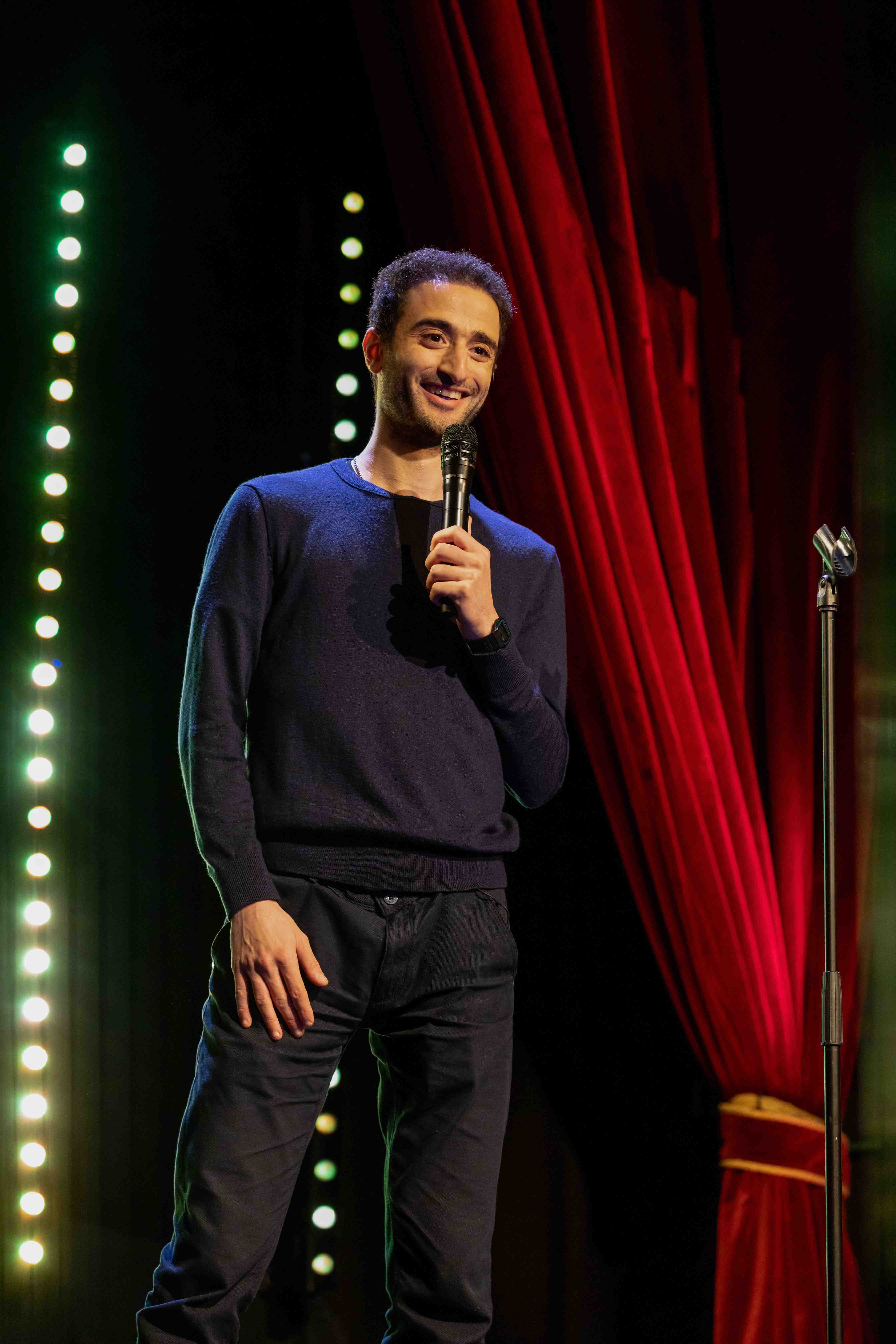 man with thick, dark, short hair wearing a dark sweater and dark jeans standing on stage, smiling while holding a microphone in front of a red curtain