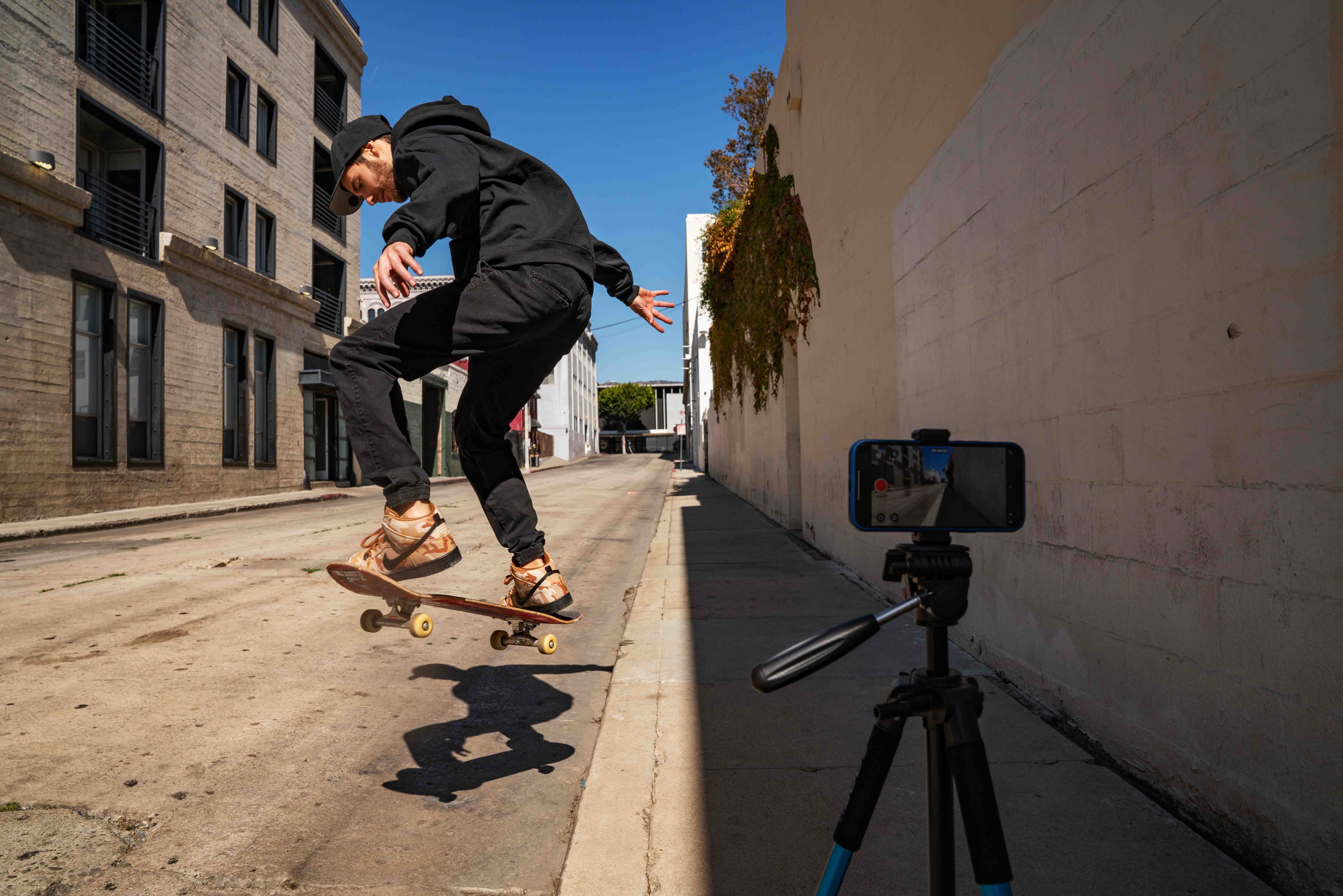 man wearing a black hat, black hoodie, black jeans and tan-camo shoes doing a trick on a skateboard on an empty street with a phone recording a video of the process