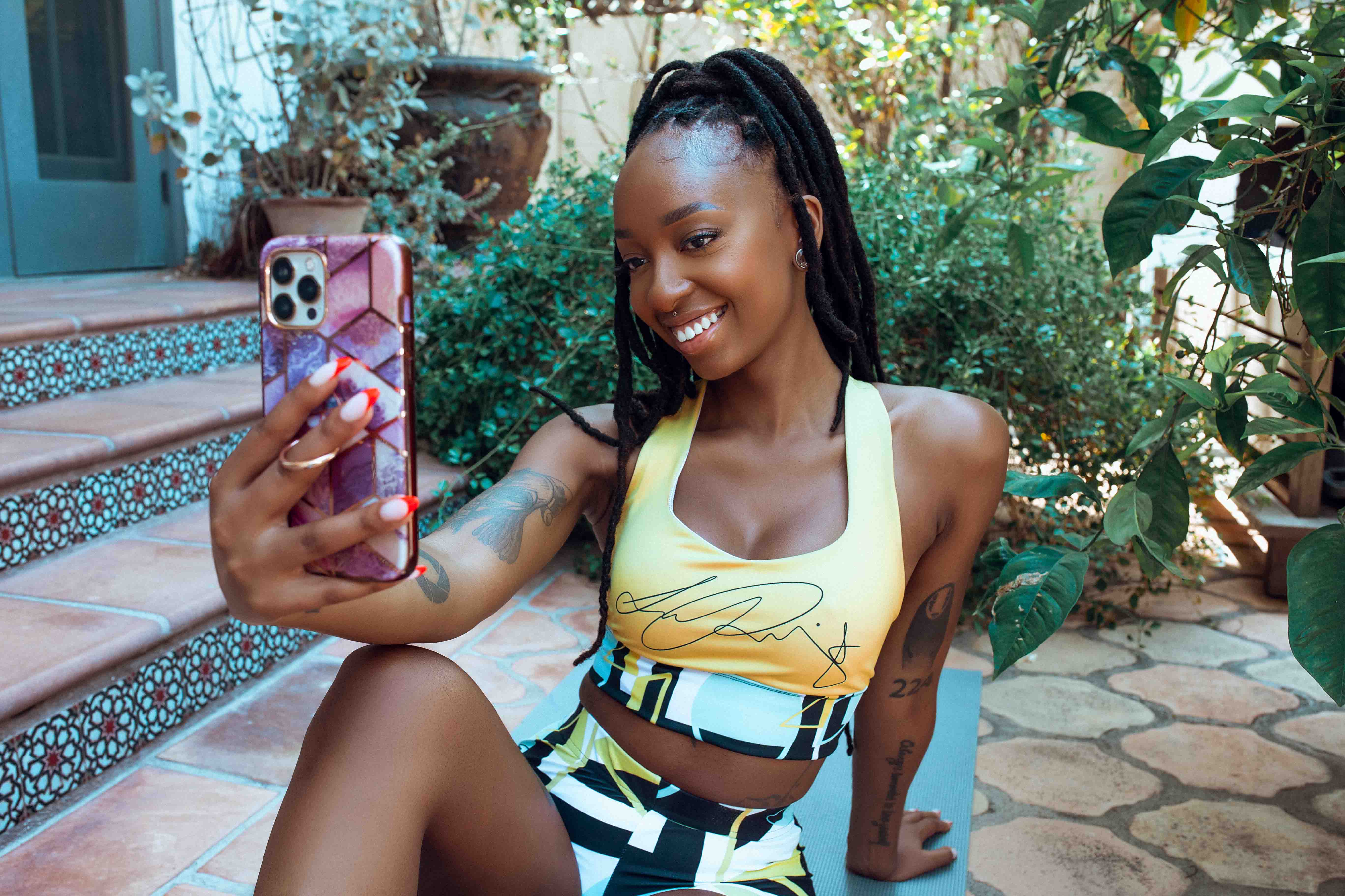 woman with long, dark cornrows pulled up into a loose ponytail, wearing a yellow sports bra with matching athletic shorts, leaning backwards on a bench posing holding her phone up in front of her to take a photo of herself in front of a botanical atmosphere next to stairs that lead to the entrance of a house