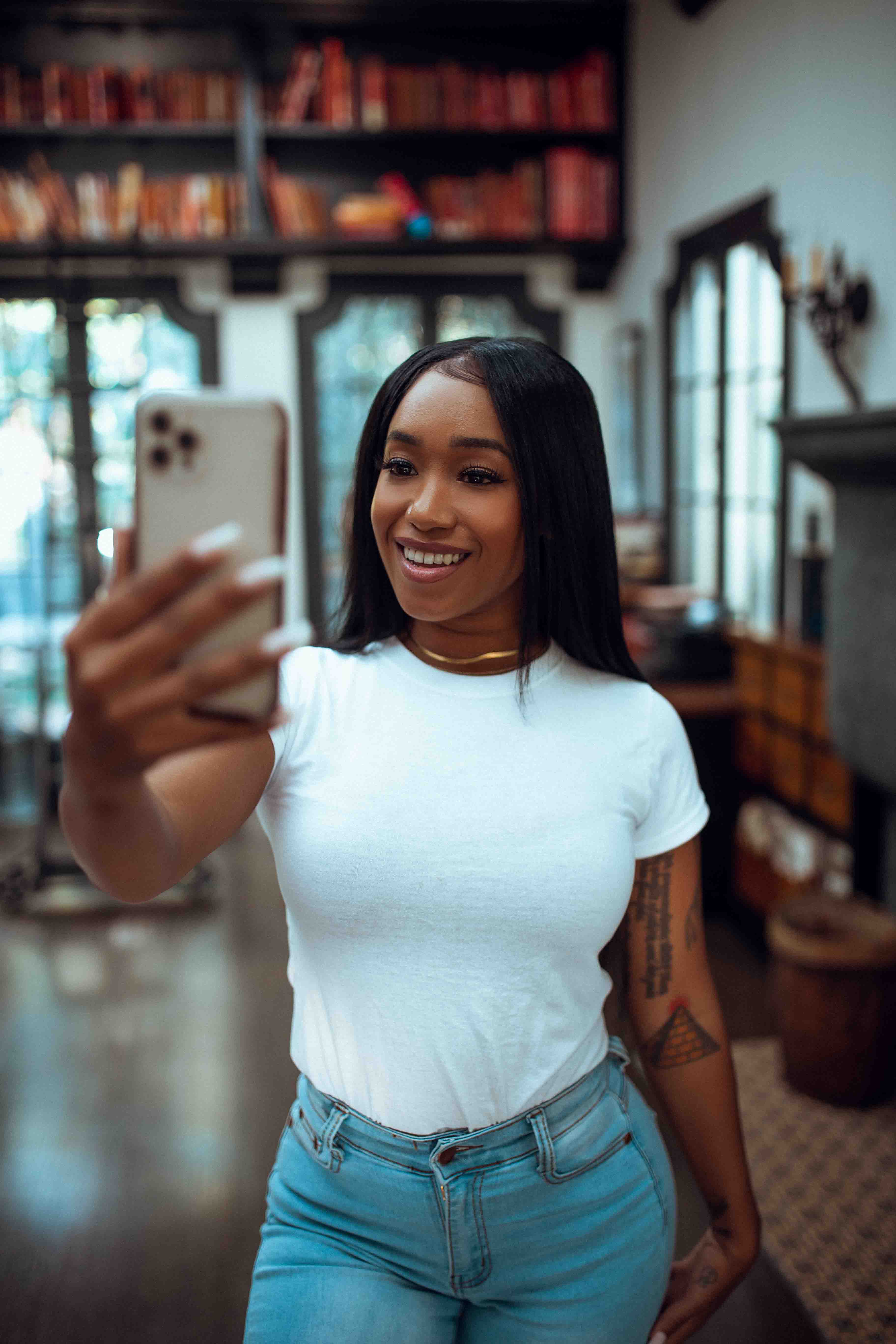 woman with dark, straight, long hair wearing a white t-shirt tucked into skinny light-wash blue jeans posing holding her phone up in front of her to take a photo of herself in front of an old library-like atmosphere