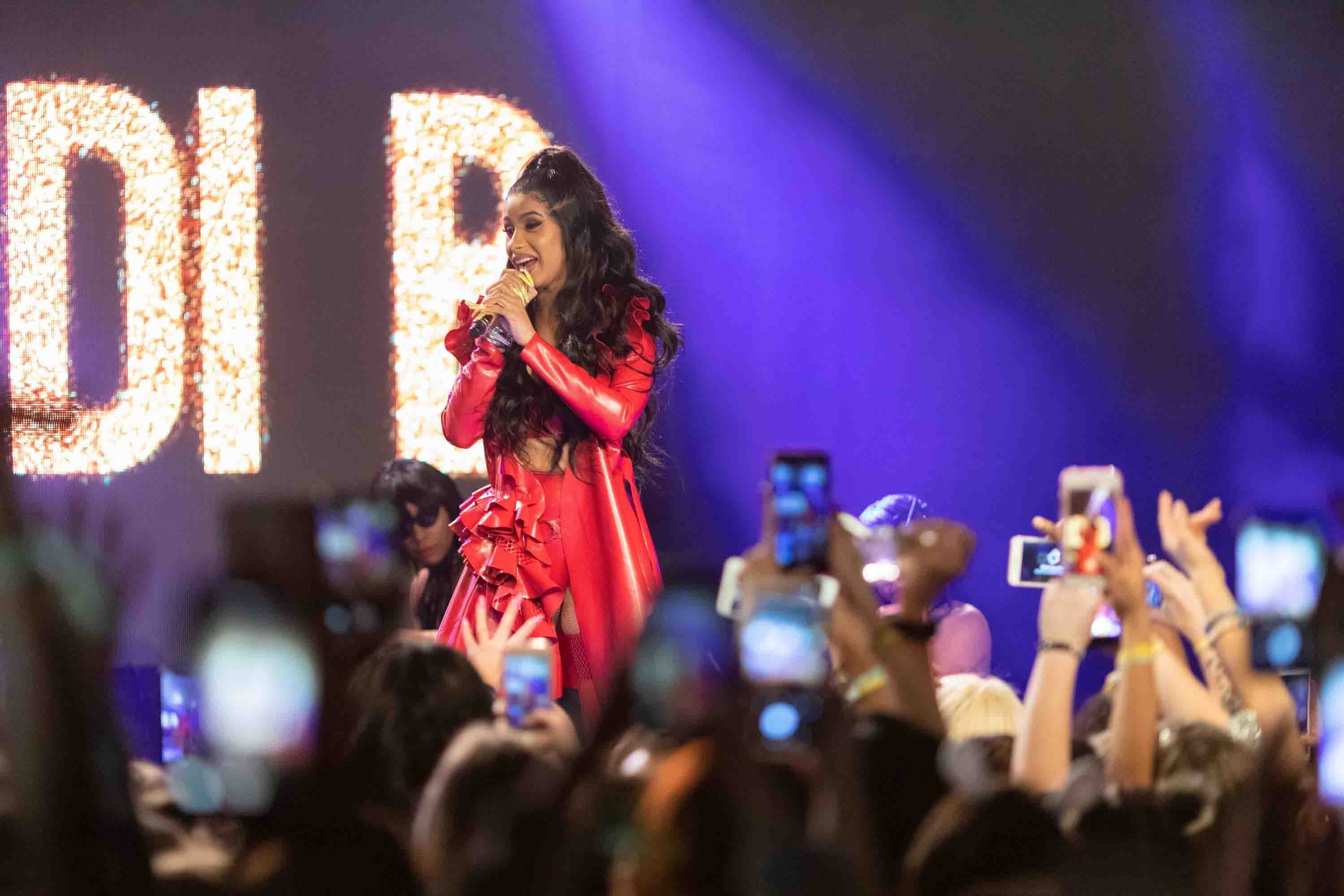 Musician, Cardi B, with dark, curly, long hair wearing a red top with matching high-waisted bottom with a red bow-like flourishment and a red glossy outer trenchcoat holding a microphone up to her open mouth on stage in front of an audience with fans recording her on their mobile phones
