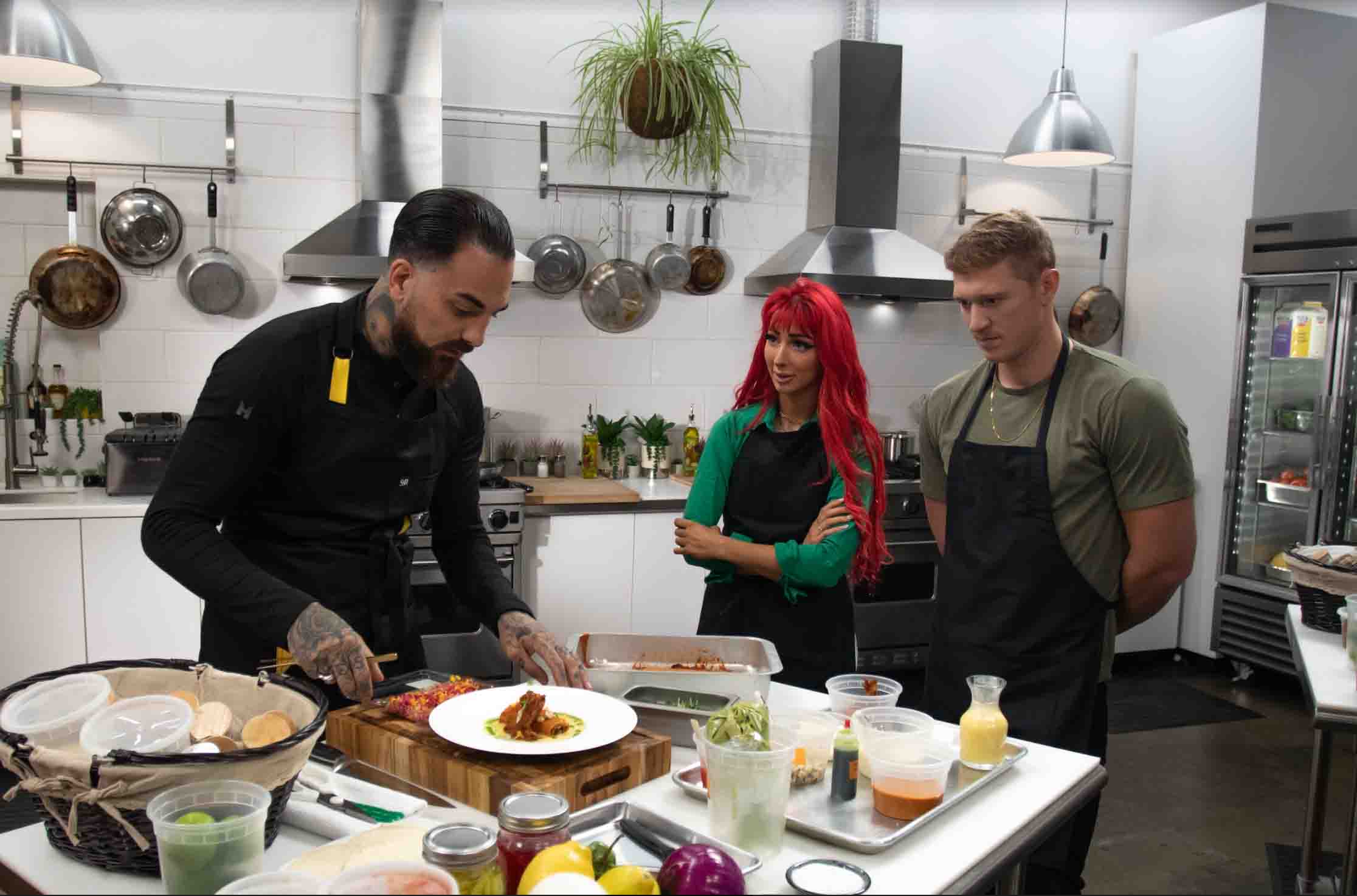 in a kitchen setting a male chef with dark slicked back hair in a black longsleeve shirt and black apron reaching out for a prepared dish to judge in front of two contestants: one woman with long dyed red hair wearing a longsleeve green shirt and black apron and one man with short dirty blonde hair wearing a short sleeve green shirt and black apron