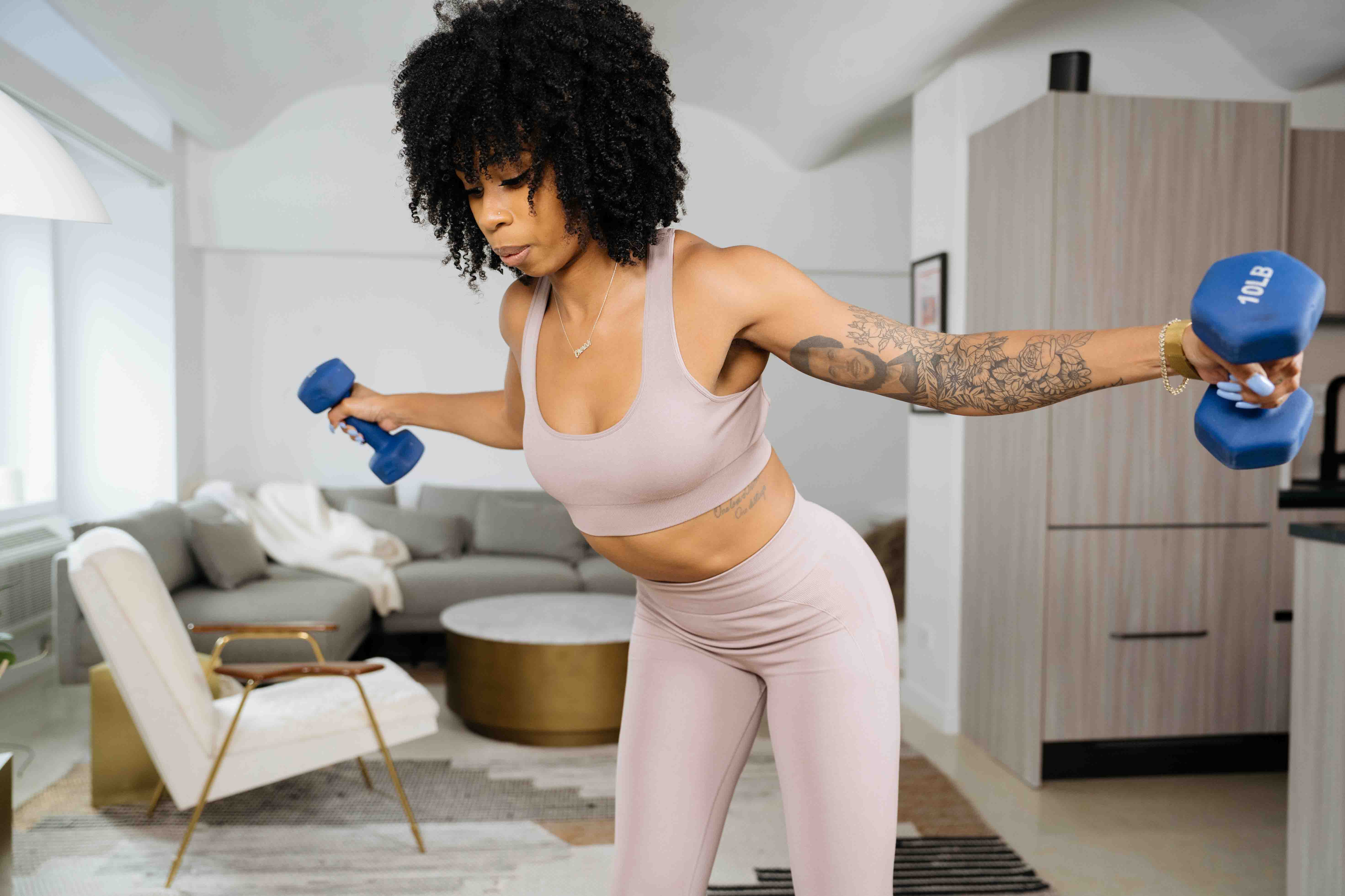 woman with dark curly hair wearing a light pink/purple sports bra with matching leggings, holding two ten pound weights outward from her body in a living room setting